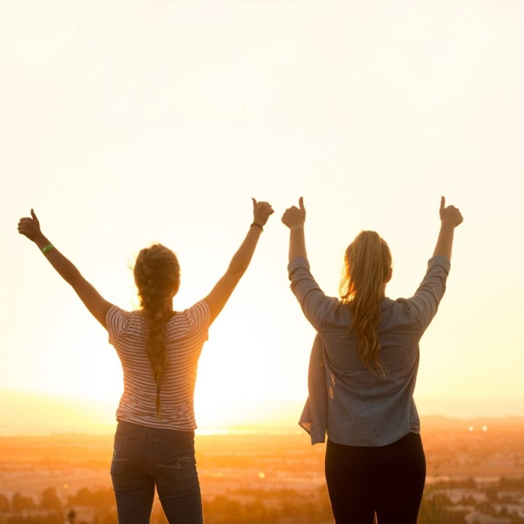 two girls looking out over the sunset with their hands in the air
